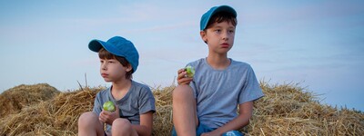 Zwei Jungen sitzen auf Heuballen, tragen graue T-Shirts und blaue Kappen, einer tr&auml;gt gr&uuml;ne Stiefel, der andere ist barfu&szlig;.