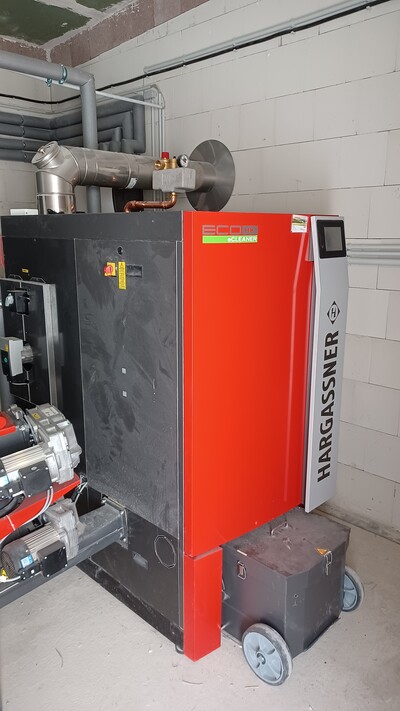 Blick in einen Heizraum mit Hackschnitzelfeuerung; die Aschekästen im Vordergrund müssen regelmäßig geleert werden. Die Anlage informiert den Betreiber rechtzeitig per App oder Anzeige darüber. - © Gerold Tammen /media/media/68541F19-DBDE-BD19-61138C143CF38A84.jpg