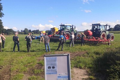 Mehrere Personen stehen auf einem Feld neben landwirtschaftlichen Maschinen und einem Informationsschild.