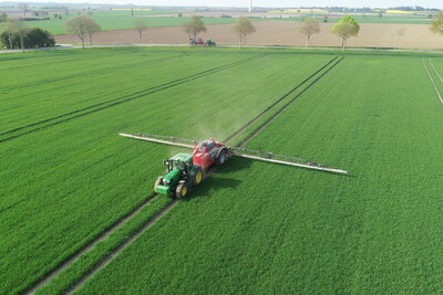 Traktor spr&uuml;ht Pflanzenschutzmittel auf ein gro&szlig;es, gr&uuml;nes Feld. Im Hintergrund sind B&auml;ume und ein Windrad zu sehen.