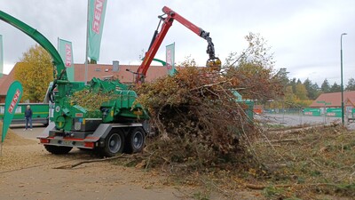Ein gro&szlig;er Holzzerkleinerer verarbeitet mit einem Greifarm &Auml;ste und Zweige auf einem Platz mit Geb&auml;uden im Hintergrund.