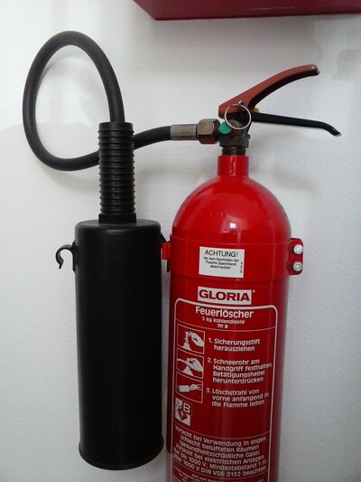 CO2-Feuerlöscher - © Thomas Middelberg Roter Feuerlöscher mit schwarzem Schlauch und Handgriff an einer weißen Wand befestigt.