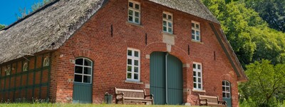- © Alfred Derks / pixabay.com Backsteinhaus mit Reetdach, grünen Türen und Fenstern. Zwei Holzbänke stehen davor, umgeben von Bäumen und blauem Himmel.