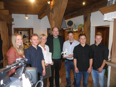 Minister Meyer (Mitte) mit daneben Sabine und Andreas Lohmüller und im Vordergrund den Kindern Alison, Luke, Devon und Jake - © Dr. Hans-Gerd Brunken Sieben Personen stehen in einem rustikalen Raum, zwei halten Urkunden und eine Skulptur.