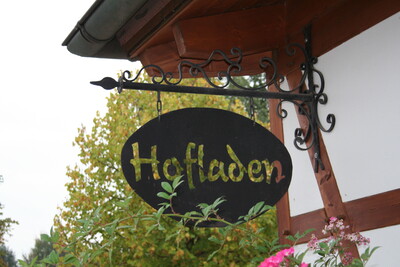 Ein ovales Schild mit der Aufschrift "Hofladen" h&auml;ngt an einem dekorativen Metallarm an einem Geb&auml;ude.