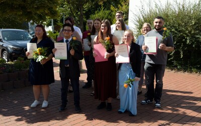 Eine Gruppe von Menschen steht drau&szlig;en auf einem Gehweg, h&auml;lt Zertifikate und Blumen in den H&auml;nden.