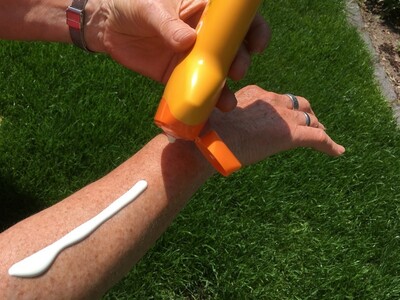 UV-Schutz ist wichtig - © Anke Fröhlich Eine Person trägt Sonnencreme aus einer gelben Flasche auf ihren Unterarm auf, während sie auf Gras steht.