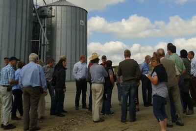 Gruppe von Menschen steht im Freien vor gro&szlig;en Silos unter bew&ouml;lktem Himmel.