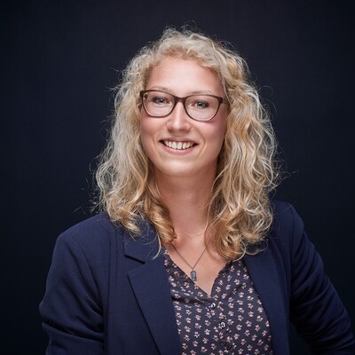 Madlen Klapproth - © Madlen Klapproth Eine Frau mit lockigem blondem Haar und Brille lächelt in die Kamera. Sie trägt ein dunkelblaues Sakko und eine gemusterte Bluse.