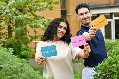 Zwei Personen stehen drau&szlig;en und halten bunte Karten mit der Aufschrift "Multitalent" in die Kamera, umgeben von gr&uuml;nen Pflanzen.
