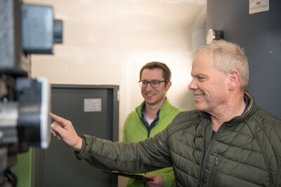 Zwei M&auml;nner in gr&uuml;nen Jacken stehen in einem Raum. Einer bedient eine Maschine, der andere h&auml;lt ein Tablet.