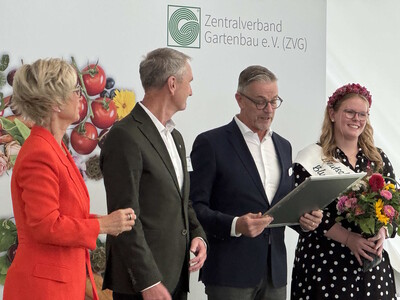 Der Vorsitzende des Bundesverbandes Zierpflanzen im ZVG Frank Werner überreicht die Urkunde - © Beate ter Hell Vier Personen stehen vor einem Banner des Zentralverbands Gartenbau. Eine Frau hält Blumen und trägt eine Schärpe.