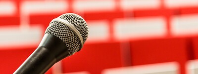 Mikrofon im Vordergrund, unscharfe rote Stuhlreihen im Hintergrund, vermutlich in einem Auditorium.