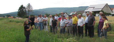 Gruppe von Menschen steht auf einer Wiese, h&ouml;rt einer Person zu. Im Hintergrund sind ein Geb&auml;ude und H&uuml;gel sichtbar.