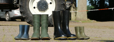 - © Gerda von Lienen Vier Paar Gummistiefel in verschiedenen Größen stehen vor einem blauen Traktor auf einem gepflasterten Platz.