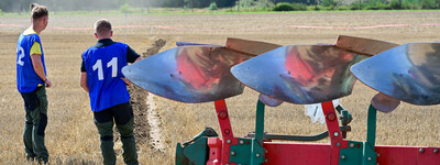 - © Andreas Teichler Zwei Personen in blauen Trikots stehen neben einem Pflug auf einem Feld.