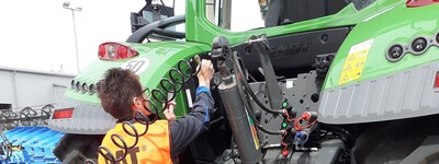 - © Eide Hauschild Person in orange Weste arbeitet an der Rückseite eines großen grünen Traktors auf gepflastertem Boden.