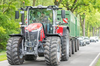 Ein roter Traktor mit gr&uuml;nem Anh&auml;nger f&auml;hrt auf einer von B&auml;umen ges&auml;umten Stra&szlig;e.