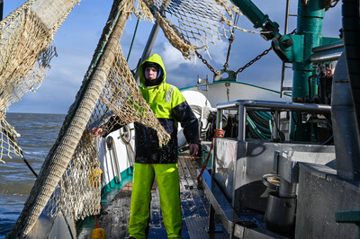 Person in gelber Regenkleidung steht auf einem Fischerboot neben Netzen, Meer im Hintergrund.