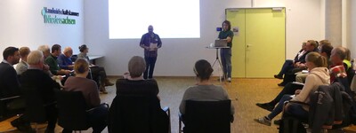 - © Wiebke Schlich Ein Vortrag in einem Konferenzraum, etwa 15 Personen sitzen im Halbkreis, ein Präsentator steht vorne.