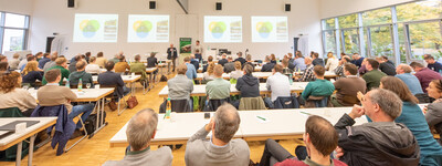 - © Wolfgang Ehrecke Viele Personen sitzen in einem großen Konferenzraum und hören einem Vortrag zu. Präsentationen sind an der Wand zu sehen.