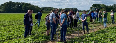 Gruppe von Menschen steht auf einem Feld unter blauem Himmel mit Wolken, neben einem wei&szlig;en Banner.