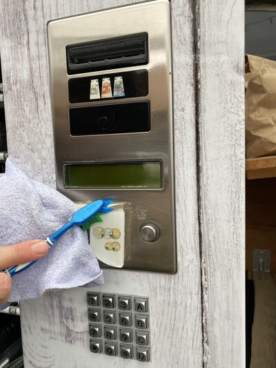 Verkaufsautomaten in der Direktvermarktung - © Julia Klöpper Eine Hand reinigt mit Zahnbürste und Tuch eine Gegensprechanlage mit Tastenfeld an einer Wand.