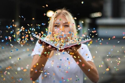 Person in wei&szlig;em T-Shirt h&auml;lt ein Buch, aus dem bunte Konfetti fliegen. Hintergrund unscharf.